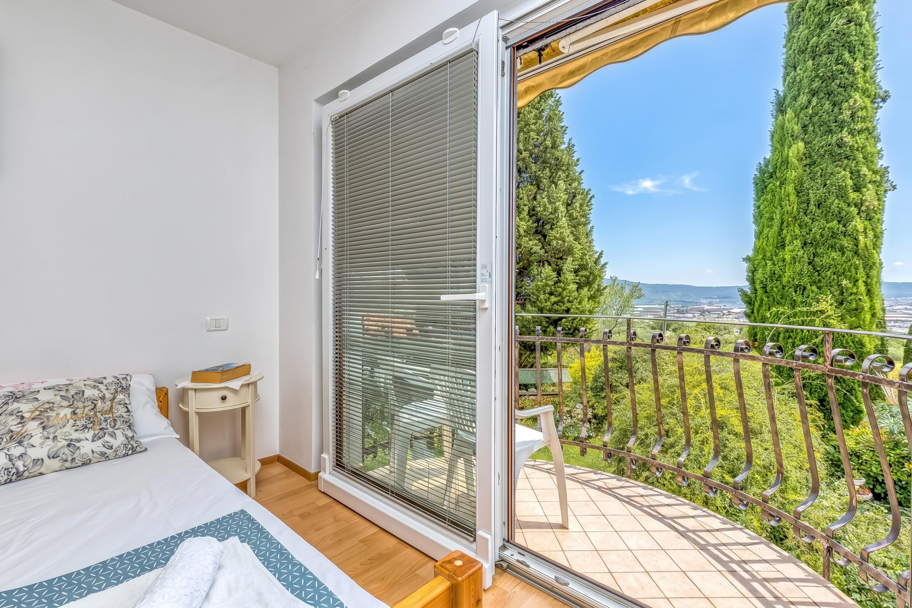 A bedroom with a bed, a nightstand, a window with blinds, and a balcony with a view of greenery and a fence.