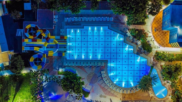 Vista aèria d'un hotel amb una gran piscina central, zones d'estar a l'aire lliure i un tobogan aquàtic.