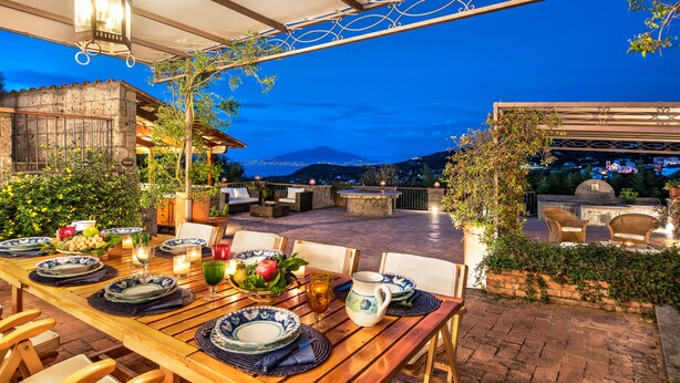 An outdoor dining area with a wooden table set for two, surrounded by chairs, under a pergola with hanging lights. - Country Lemonvilla (Massa Lubrense)