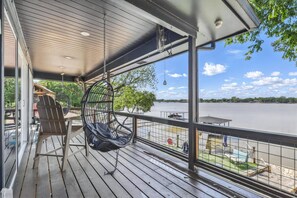 A wooden deck with a hanging chair, a table, and a view of a lake. - Waterfront Getaway on Lake Granbury! Pet Friendly, Lake Front with Boat House and Hot Tub! (Granbury)