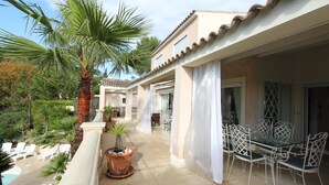 Eine überdachte Terrasse im Freien mit Pool, Palmen und einer Essecke mit weißen Stühlen und einem Glastisch.