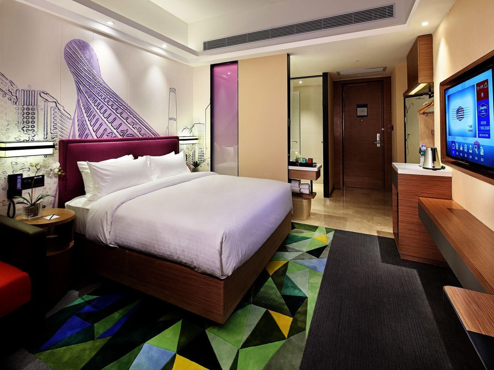 A modern hotel room with a large bed, a desk, a TV, and a bathroom visible through an open door.