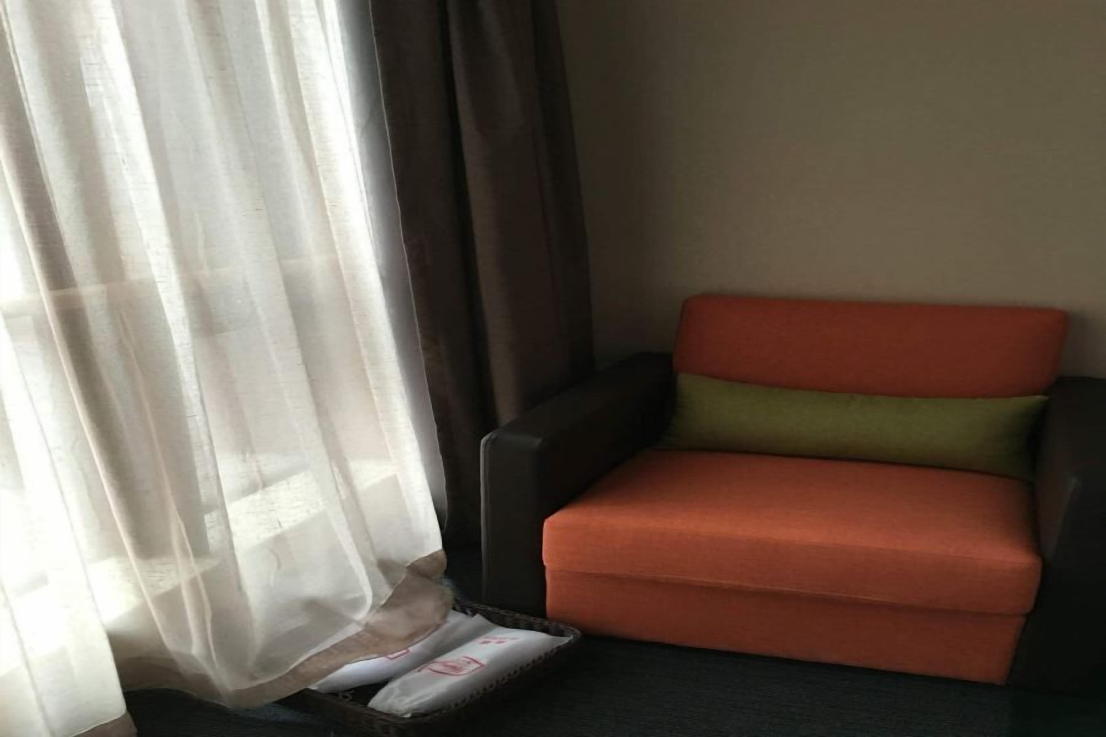 A room with a single orange chair, a black armchair, and a window with sheer curtains.