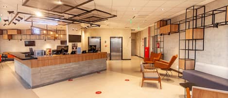A modern hotel lobby with a reception desk, seating area, elevators, and a bar.
