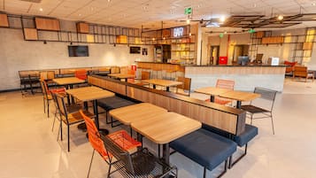 Una cafetería moderna con mesas de madera y sillas naranjas, una zona de bar y una sección de asientos con bancos azules.