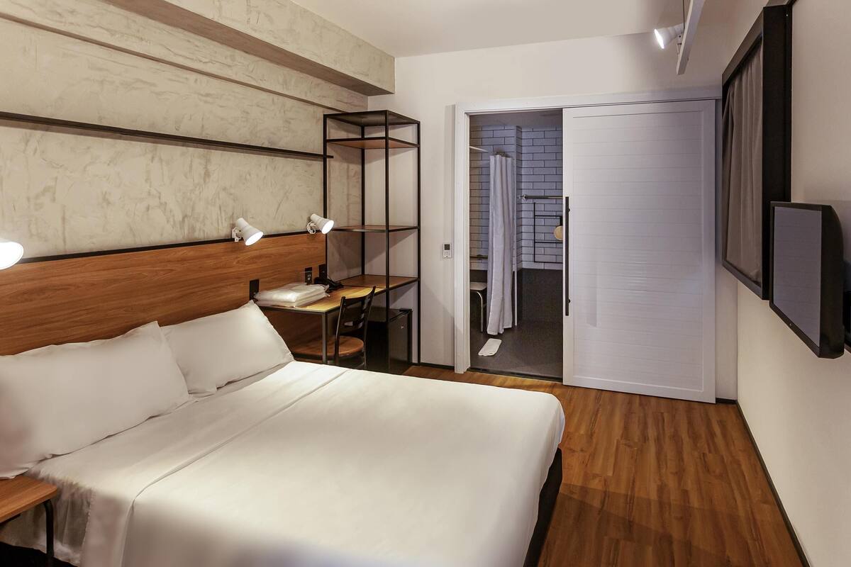 A hotel room with a bed, bedside table, lamp, and a TV mounted on the wall.