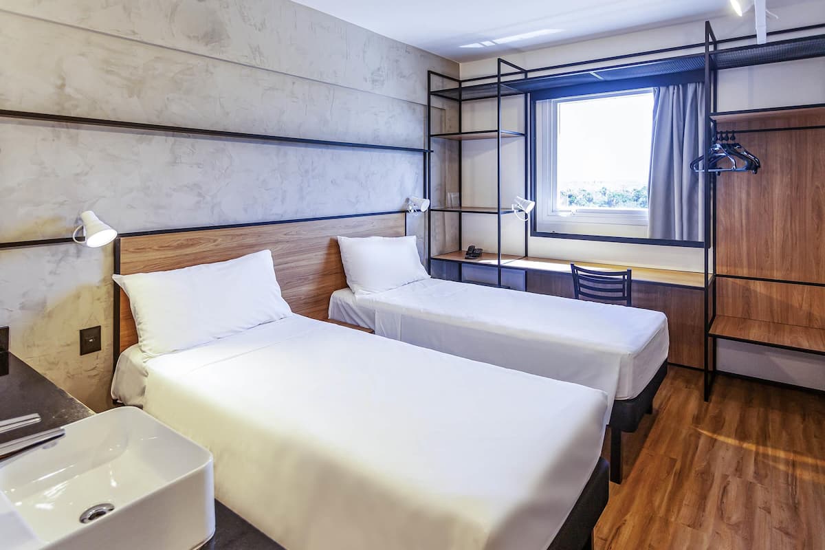 A hotel room with two beds, a desk, a chair, a window with curtains, and a wardrobe.