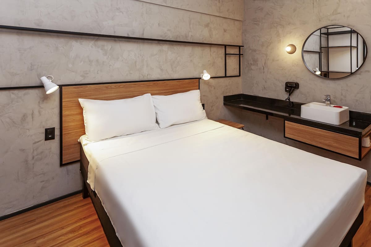 A modern hotel room with a large bed, bedside lamps, a round mirror, a wooden nightstand, and a built-in shelf with a sink.