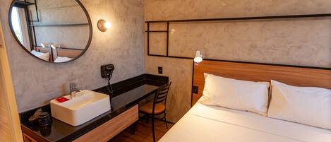 A hotel room with a wooden bed, a round mirror, a white sink, and a wooden desk.