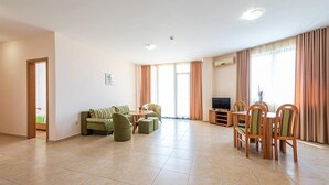 A spacious living room with a sofa set, a dining table with chairs, a television, and large windows with curtains.