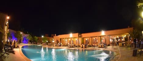 A nighttime swimming pool area with a covered seating area and a building with multiple windows and doors.