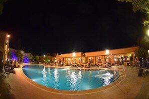 Un área de piscina nocturna con una zona de estar cubierta y un edificio con múltiples ventanas y puertas.