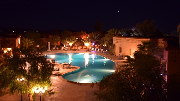 A nighttime view of a swimming pool surrounded by palm trees and lounge chairs, with a building in the background. - Palms Hotel Club (Aarab Sebbah Ziz)