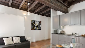 Een moderne woonkamer met een bank, een glazen eettafel met borden en bestek, een keuken met grijze kasten en een plafond met zichtbare balken.