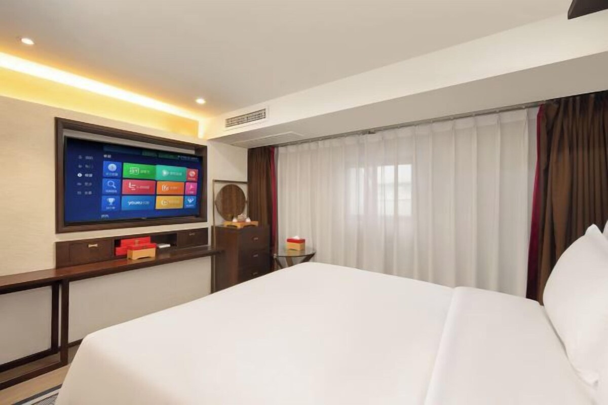 A hotel room with a large bed, a flat-screen TV mounted on the wall, a small table with a lamp, and a window with curtains.