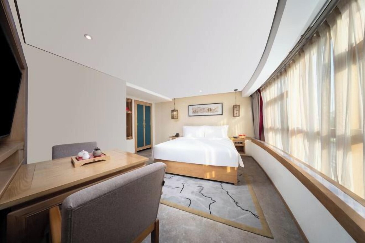 A modern hotel room with a large bed, a dining table, and a view of the city through large windows.