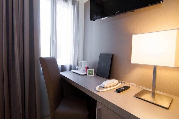 Habitación de hotel con escritorio, televisor de pantalla plana montado en la pared, lámpara de noche, teléfono y cafetera.