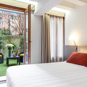 A hotel room with a bed, a small table with a vase and a plant, a chair, and a view of the outside. - Leone Hotel (Venice)