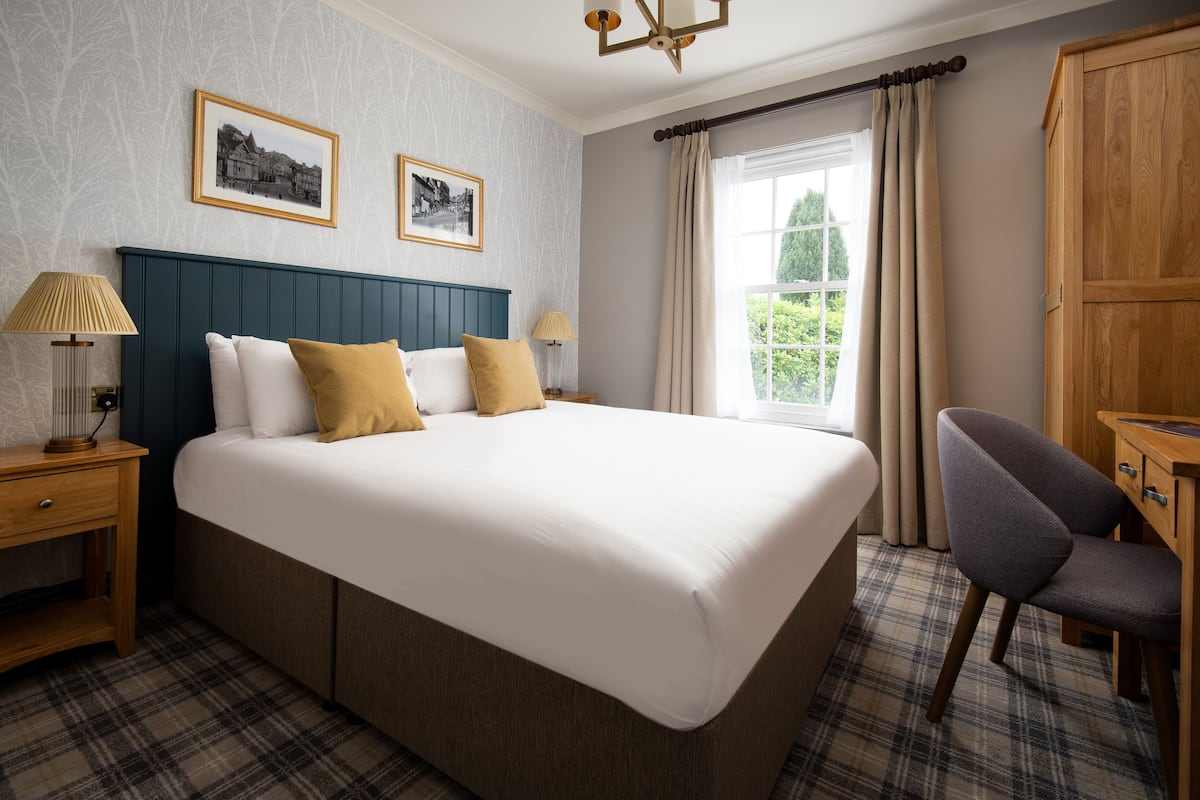 A large bed with white linens and two pillows, flanked by wooden nightstands with lamps. A plaid-patterned carpet covers the floor, and a wooden wardrobe is visible to the right.