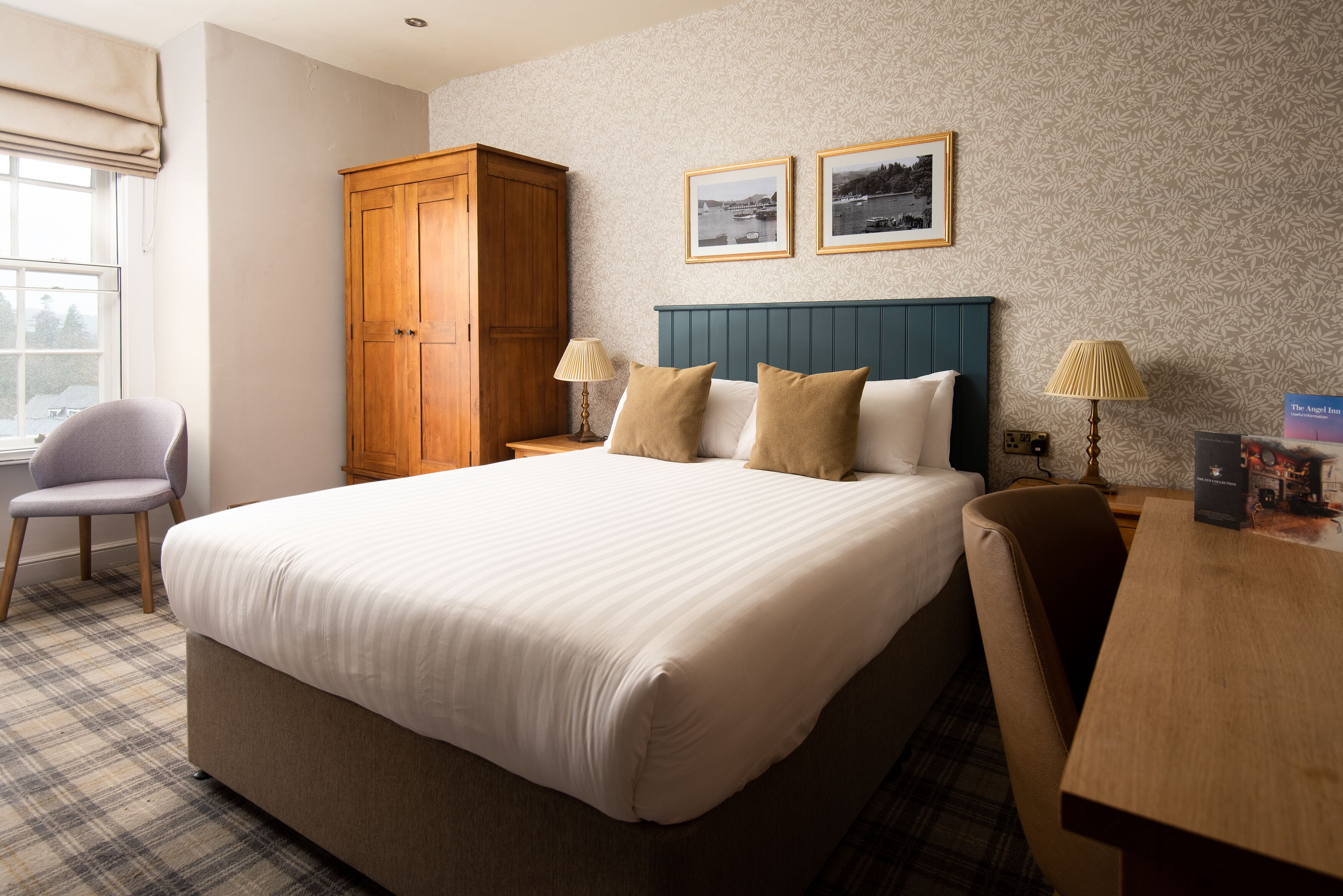 Une chambre d’hôtel avec un grand lit, une armoire en bois, deux lampes de chevet, un tapis à motifs écossais et deux tableaux encadrés au mur.