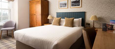 Habitación de hotel con una cama grande, un armario de madera, dos lámparas de noche, una alfombra estampada de cuadros y dos cuadros enmarcados en la pared.