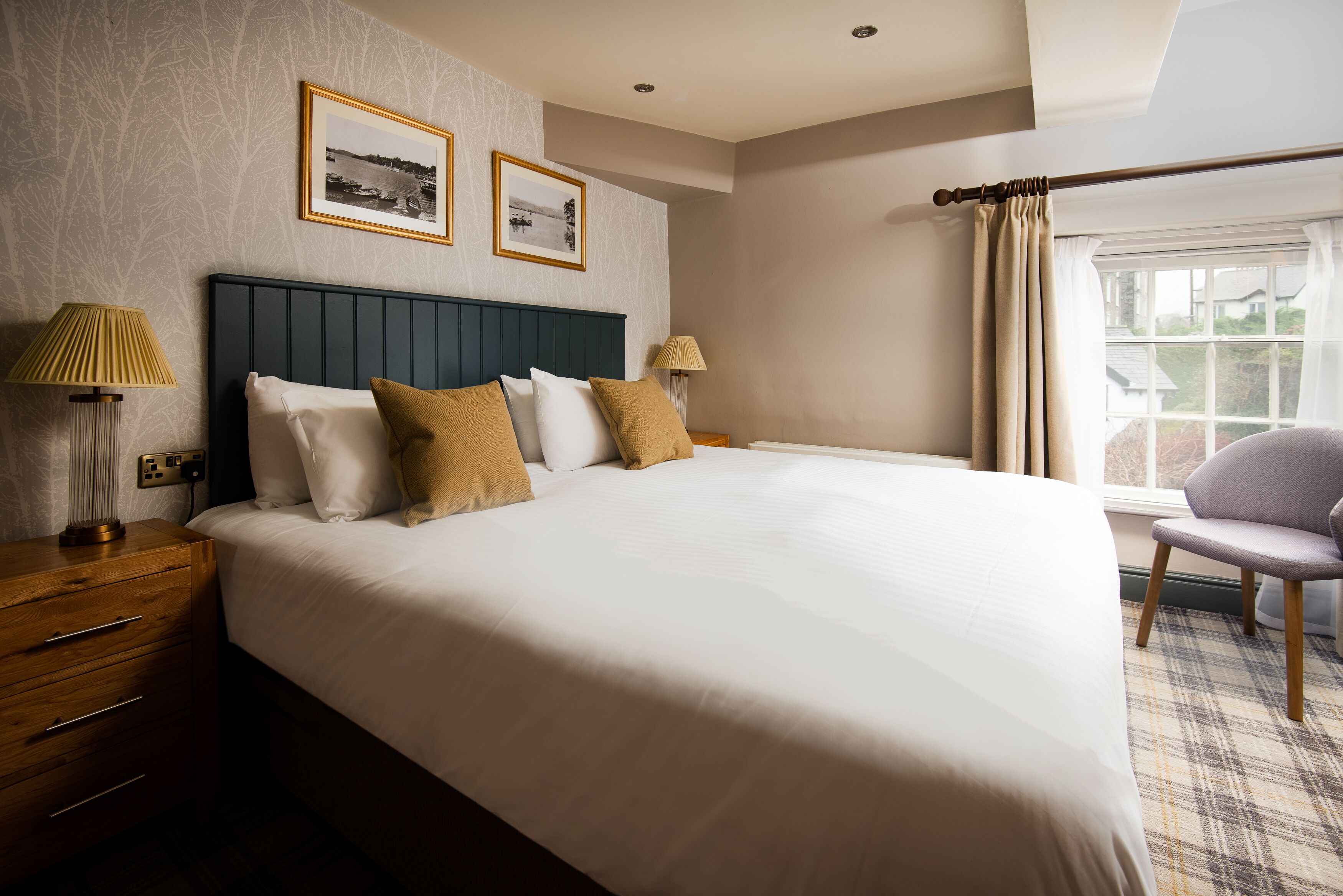 A neatly made bed with a dark headboard, two bedside lamps, and a chair by the window.