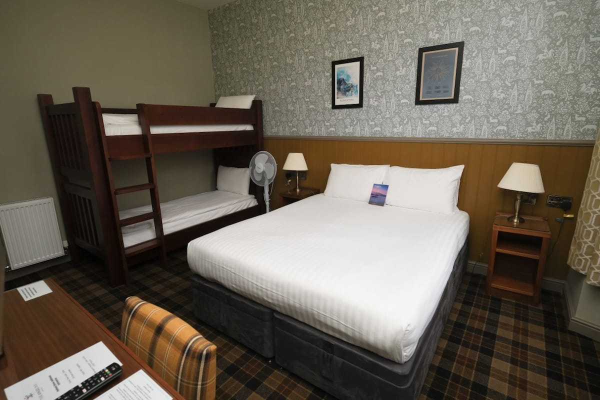 A hotel room with a bunk bed, a single bed, a desk with a chair, a bedside table with a lamp, and a wall with two framed pictures.