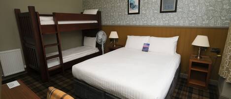 Una habitación de hotel con una cama litera, una cama individual, un escritorio con una silla, una mesita de noche con una lámpara, y una pared decorada con dos cuadros enmarcados.