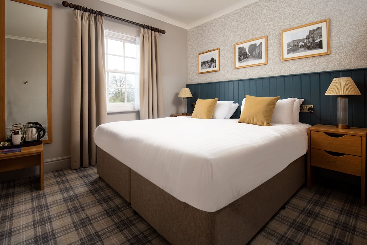 A hotel room with a large bed, a wooden nightstand, a bedside table with a lamp, and a window with curtains. There are three framed pictures on the wall above the bed.