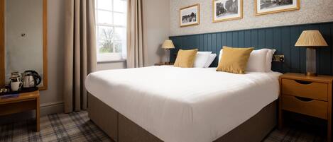 A hotel room with a large bed, a wooden nightstand, a bedside table with a lamp, and a window with curtains. There are three framed pictures on the wall above the bed.