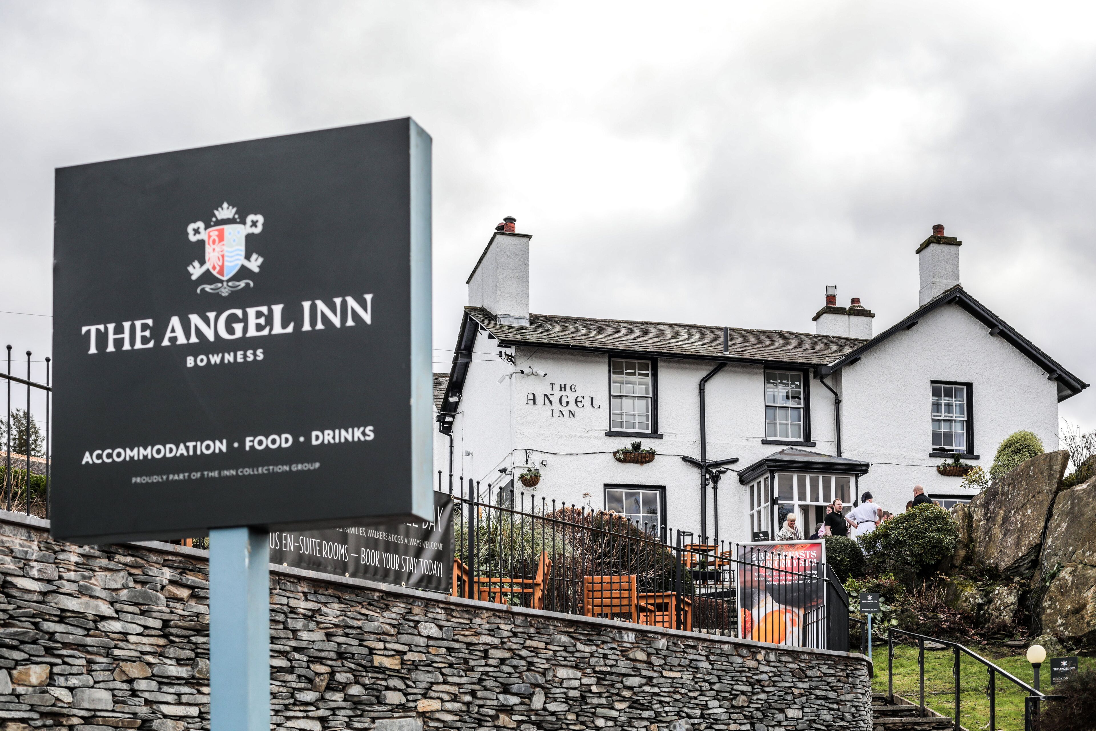 A sign for The Angel Inn in Bowness, featuring a stone wall and a white building with black trim.