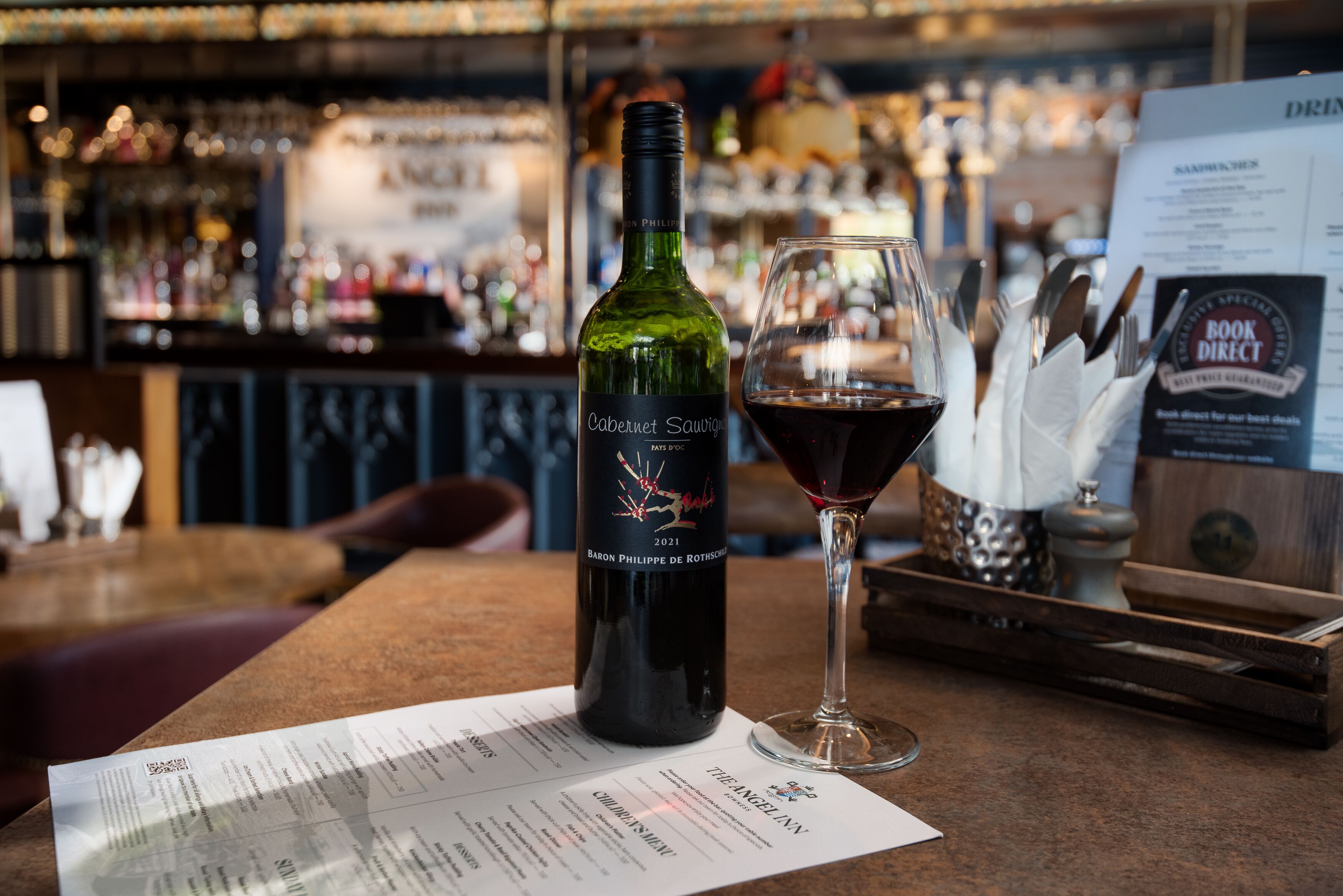 A bottle of 2021 Baron Philippe de Rothschild Cabernet Sauvignon wine and a glass of red wine on a bar counter with a menu in front.