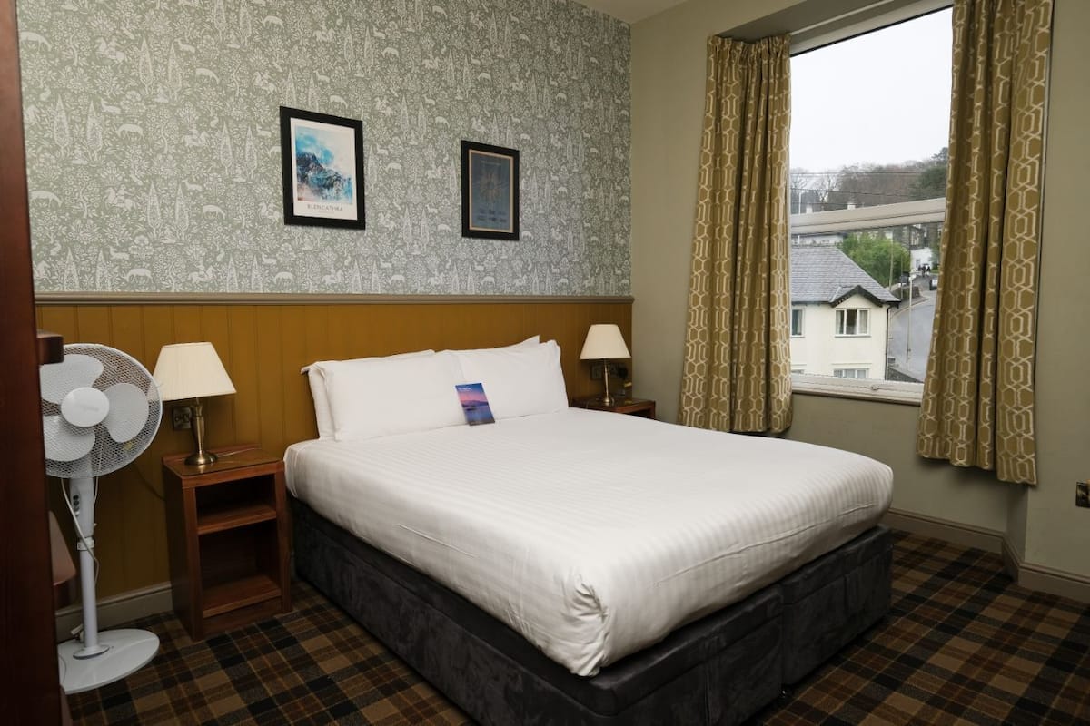 A hotel room with a bed, two bedside tables, a standing fan, and a window with curtains.