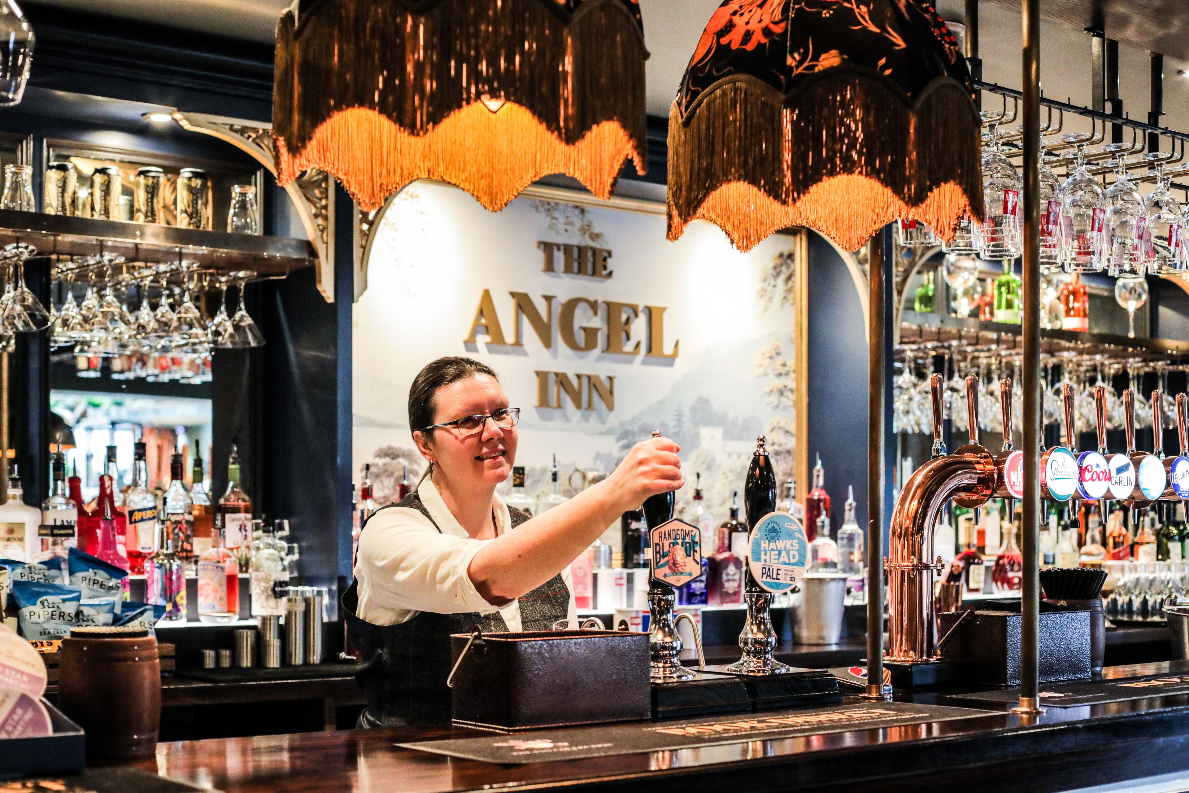 Un'area bar con un barista, un cartello con la scritta "The Angel Inn" e una selezione di bottiglie e bicchieri.