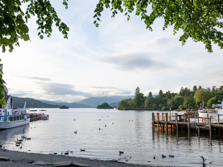A calm lake with boats docked at a pier, surrounded by trees and mountains in the background.