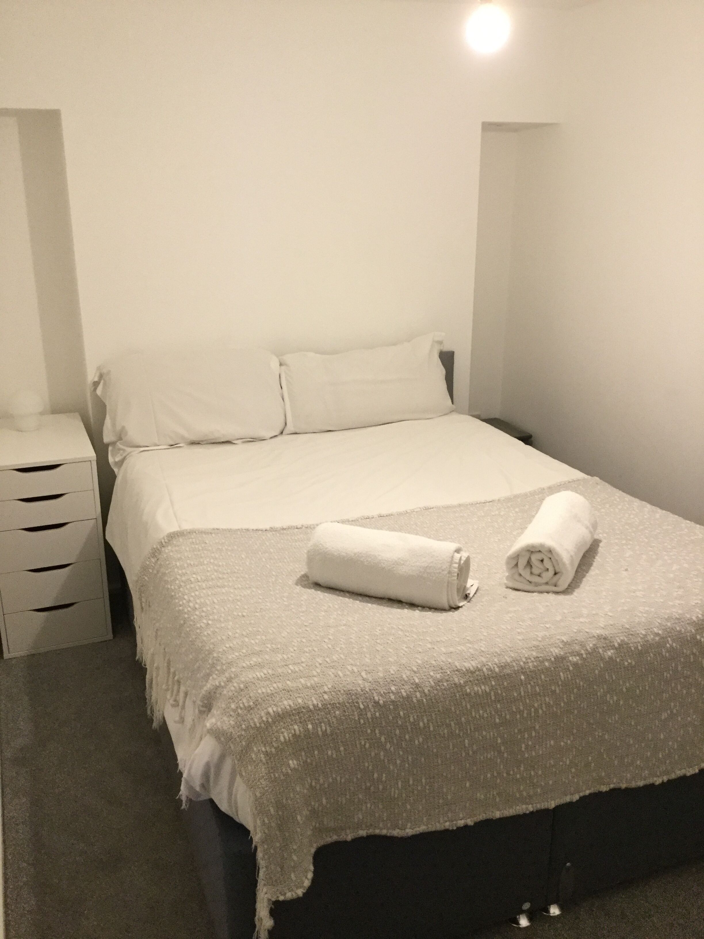 Een net opgemaakt bed met een opgerolde handdoek, een nachtkastje en een effen wand met een lamp.