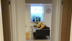 Un pasillo que conduce a una sala de estar con un sofá gris, un cojín amarillo, una ventana con una planta y un reloj en la pared.