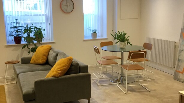 Una sala de estar moderna con un sofá gris, cojines amarillos, una mesa de comedor redonda con cuatro sillas y dos ventanas con plantas en macetas.