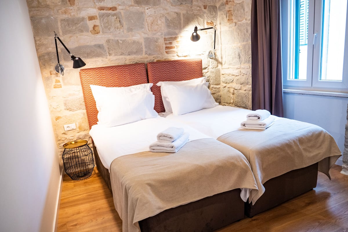 A double bed with a stone wall backdrop, two bedside lamps, and a window with curtains.
