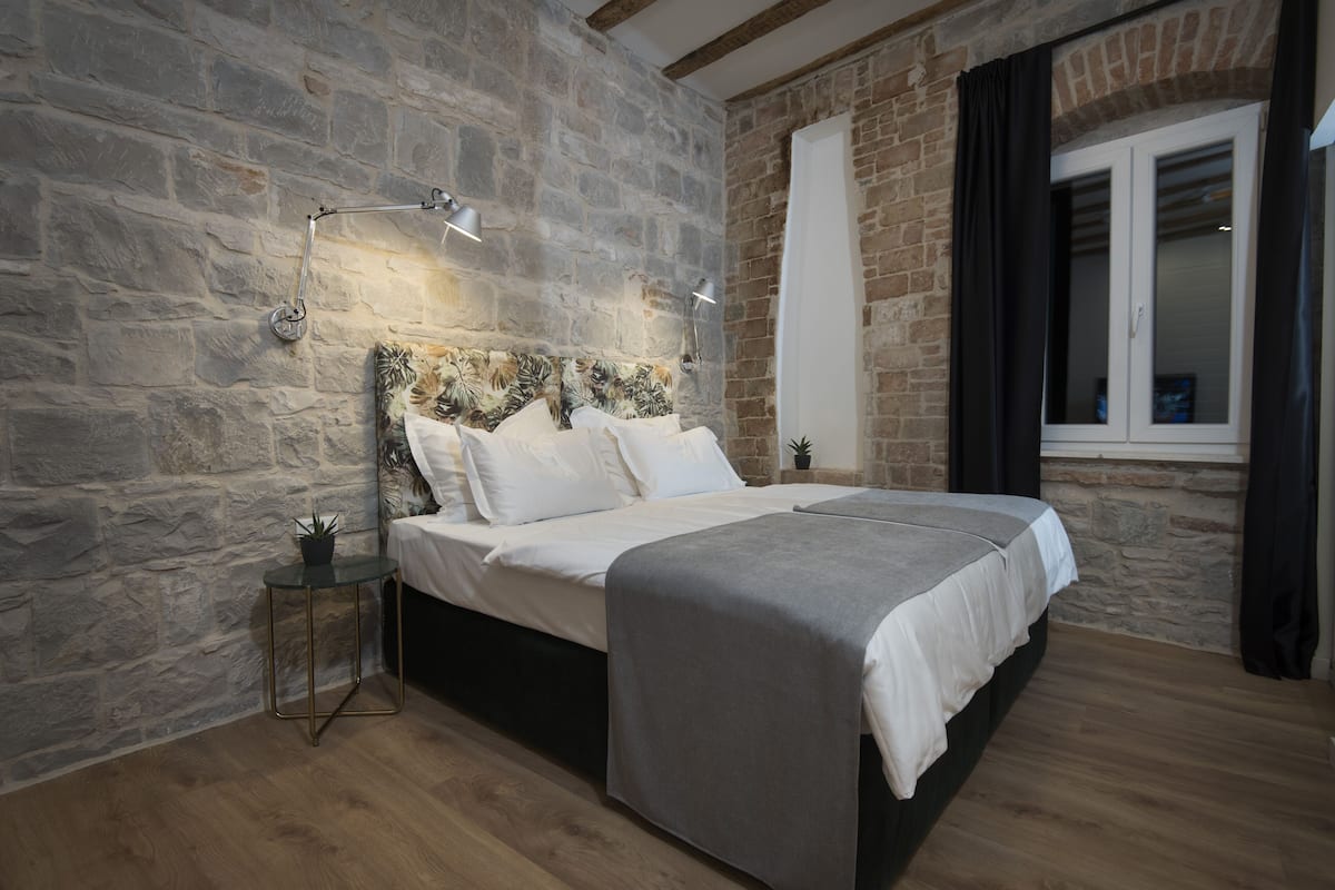 A bedroom with a stone wall, a bed with white bedding, a small side table, and a window with curtains.
