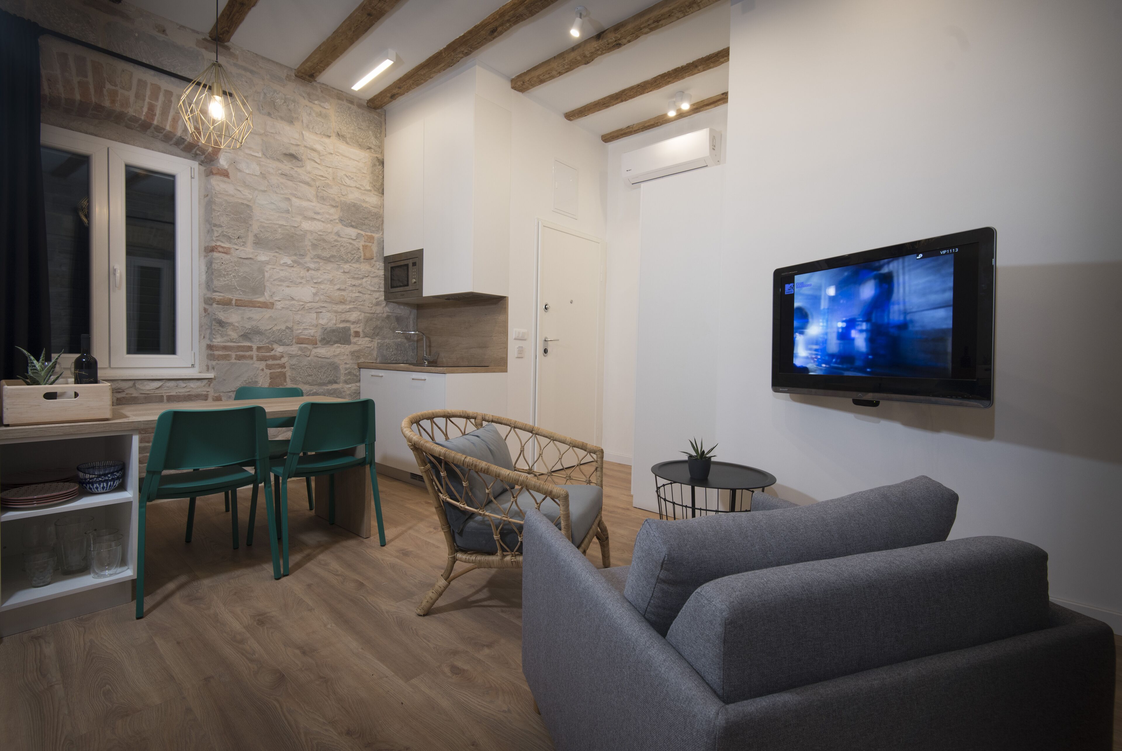A modern living room with a grey sofa, a wicker chair, and a dining area with a table and chairs. A flat-screen TV is mounted on the wall.