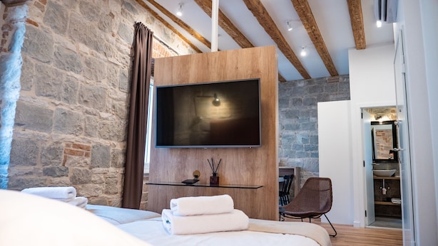 A modern bedroom with a large bed, a mounted TV, stone walls, and a wooden ceiling. - Emarconi history&luxury in Split center (Split)