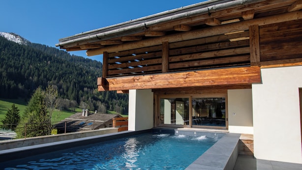 A modern pool area with a wooden pergola overhead, a clear view of a mountainous landscape, and a pool with a tiled edge. - Chalet Anthocyane, 14 pers, piscine chauffée, sauna, pistes à 100 m (LA CLUSAZ)