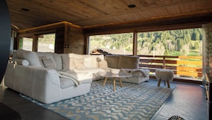 A spacious living area with a large sofa, a coffee table, and a view of a mountainous landscape through large windows. - Chalet Anthocyane, 14 pers, piscine chauffée, sauna, pistes à 100 m (LA CLUSAZ)