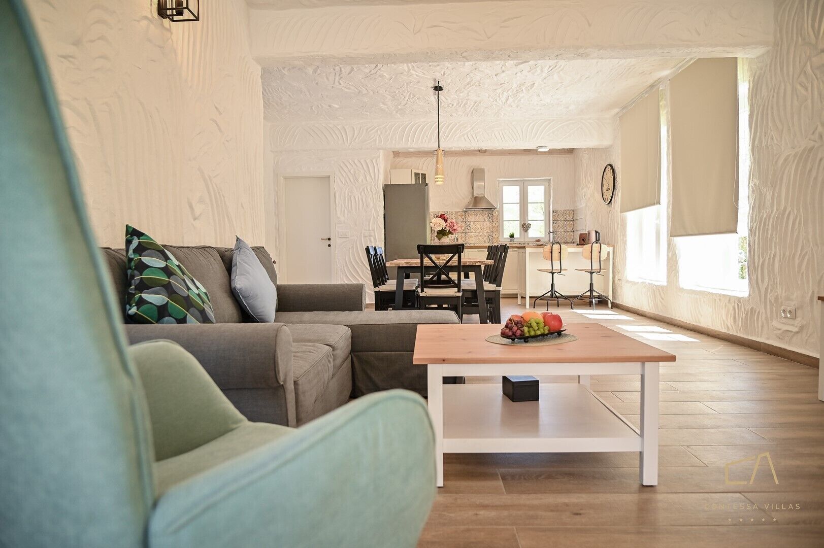 A spacious living room with a sectional sofa, a wooden coffee table with fruit, and a dining area with chairs and a kitchen in the background.