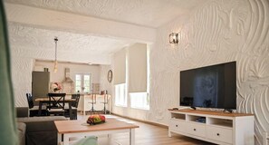 Un salon moderne avec une table basse en bois, un téléviseur posé sur un meuble blanc et un coin repas comprenant des chaises et une table.