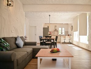 Ein modernes Wohnzimmer mit einem Ecksofa, einem Couchtisch mit Obst, einem Essbereich mit Stühlen und einer Küche im Hintergrund.