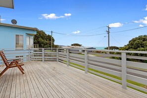 A wooden deck with a white railing, a single wooden chair, and a building with a satellite dish in the background. - Oceans 61 Beachside Charm Ocean Views & BBQ (APOLLO BAY)