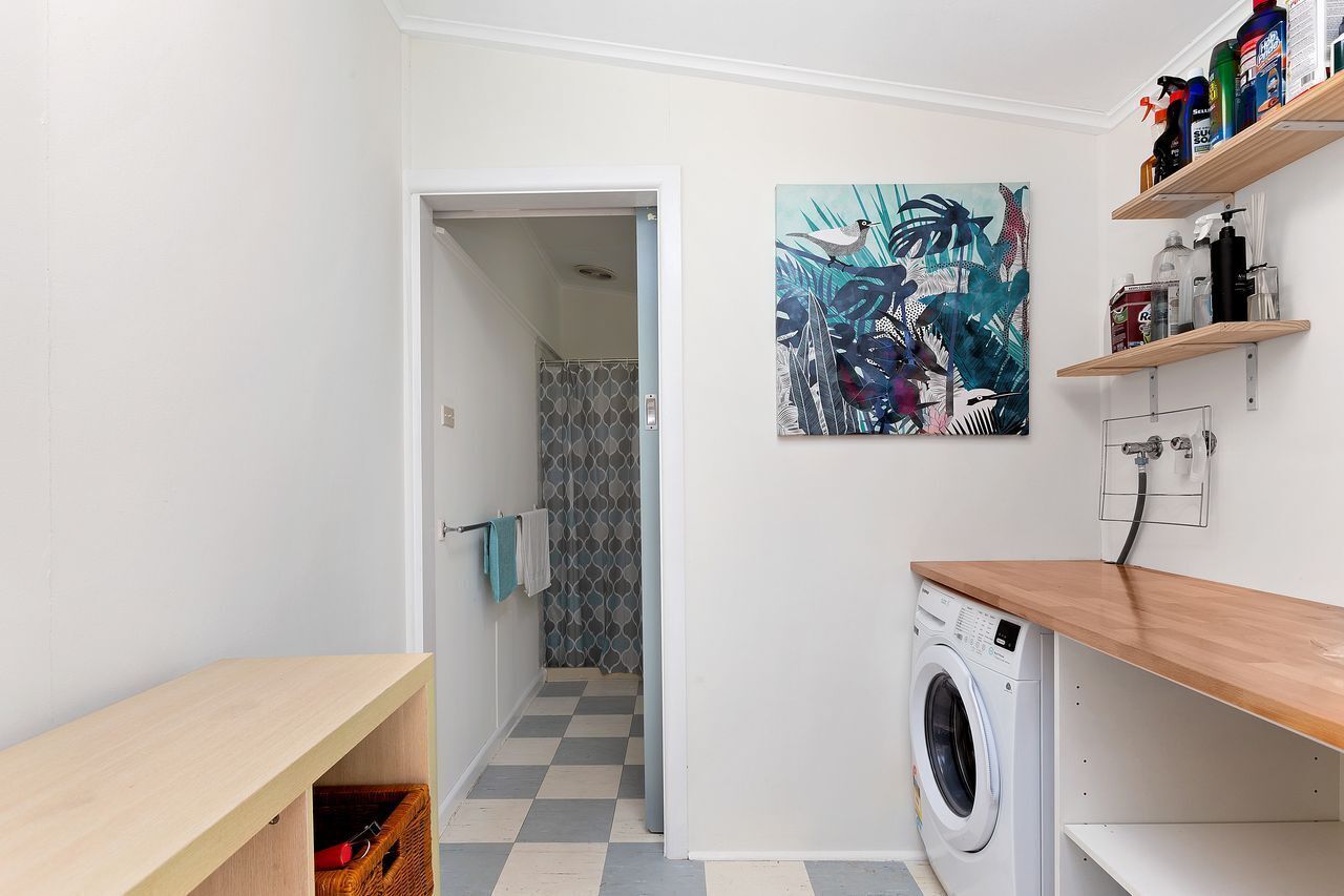 Una sala de bugaderia amb una rentadora, una prestatgeria de fusta amb productes de neteja i una porta que dona a un bany amb rajoles decoratives.