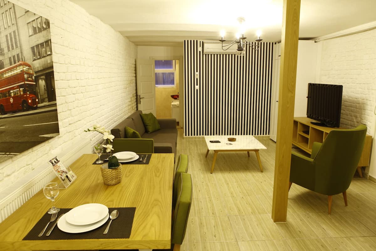 Un comedor con una mesa de madera preparada para dos comensales, un sofá verde y un televisor en una esquina.